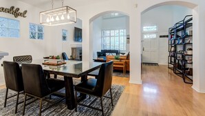 A spacious dining area with a glass-top table and black chairs, a modern chandelier, and a bookshelf filled with books and decorative items. - Located in downtown O'side - Blue Jay by the Pier (Oceanside)