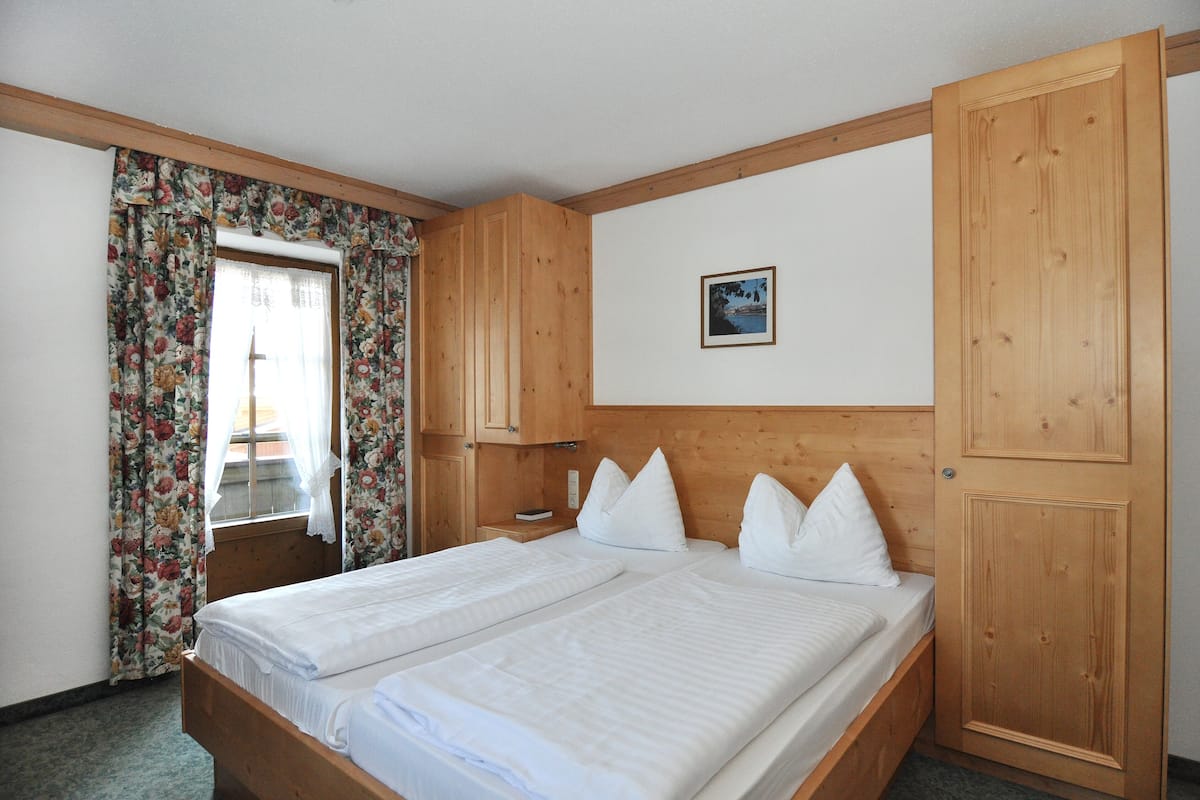 Een tweepersoonsbed met houten hoofdbord, een houten garderobe en een raam met bloemen gordijnen.