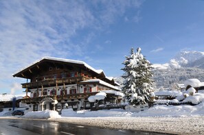 Um hotel alpino tradicional com fachada coberta de neve, uma varanda saliente e uma árvore carregada de neve à frente.