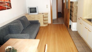 Un espacio habitable compacto con un sofá gris, una mesa de comedor de madera, un televisor sobre un mueble de madera y una zona de cocina con armarios y suelo de baldosas.