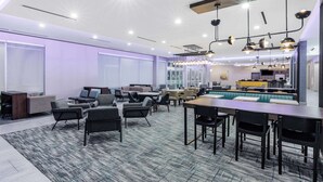 Um hall de hotel moderno com diversas áreas de estar, incluindo sofás e mesas, e um espaço de bar com banquetas.
