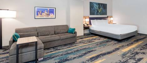 A hotel room with a gray sofa, a small side table, a bed with white bedding, a nightstand with a lamp, and a framed picture on the wall.