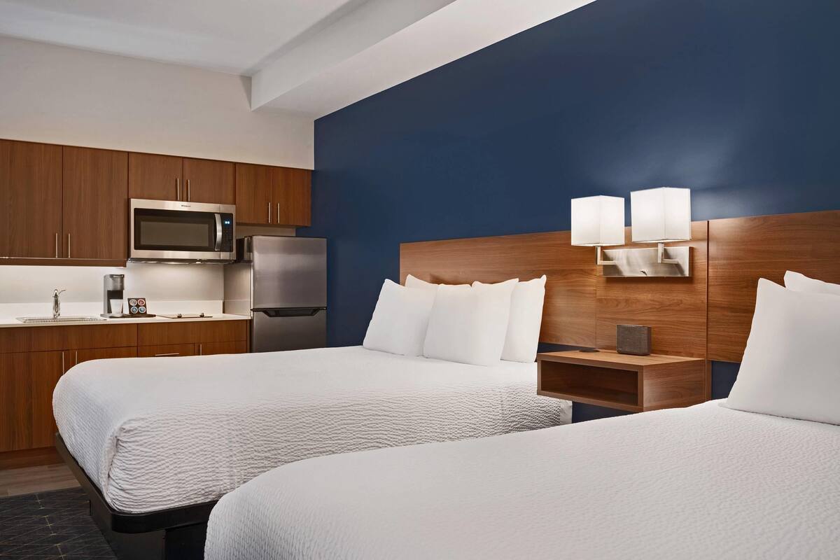 A hotel room with two beds, a kitchenette with a microwave and refrigerator, and a nightstand with lamps.