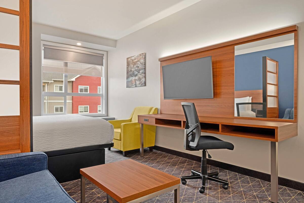 A modern hotel room with a bed, a desk, a chair, a small table, a TV, and a window with a view of buildings.