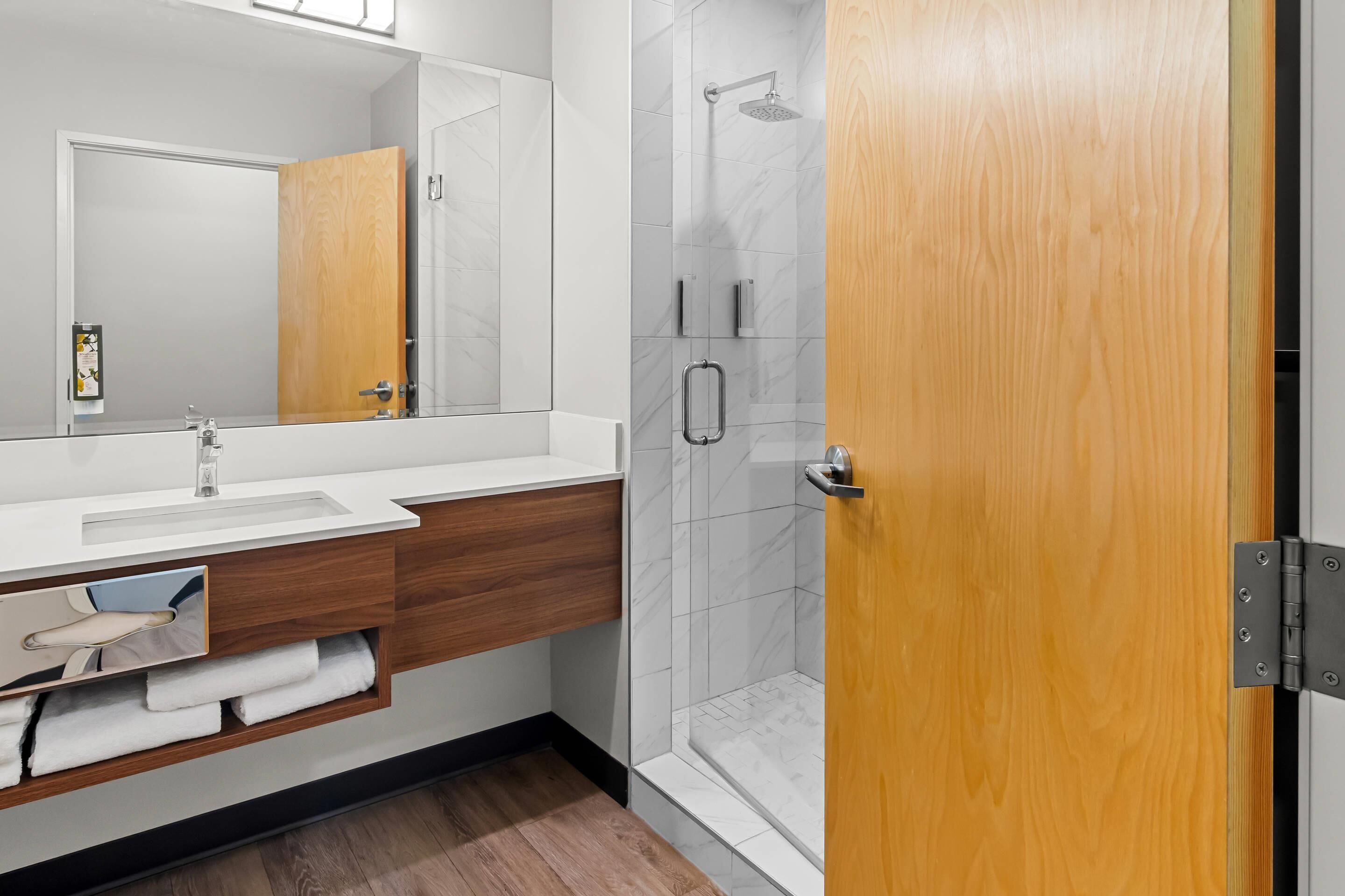 Une salle de bain moderne avec un meuble-lavabo en bois, un grand miroir, un espace douche et une porte en bois.