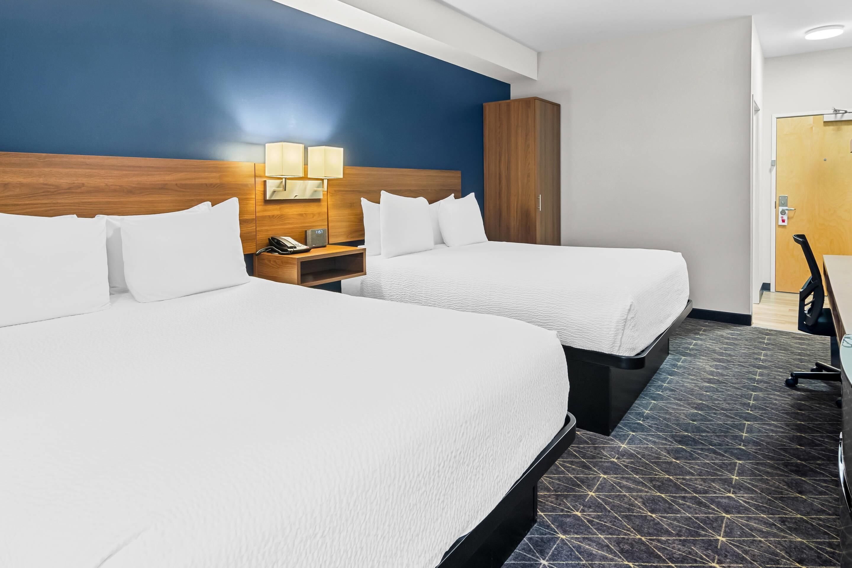 Habitación de hotel con dos camas, cabecero de madera, mesita de noche con lámpara y armario.