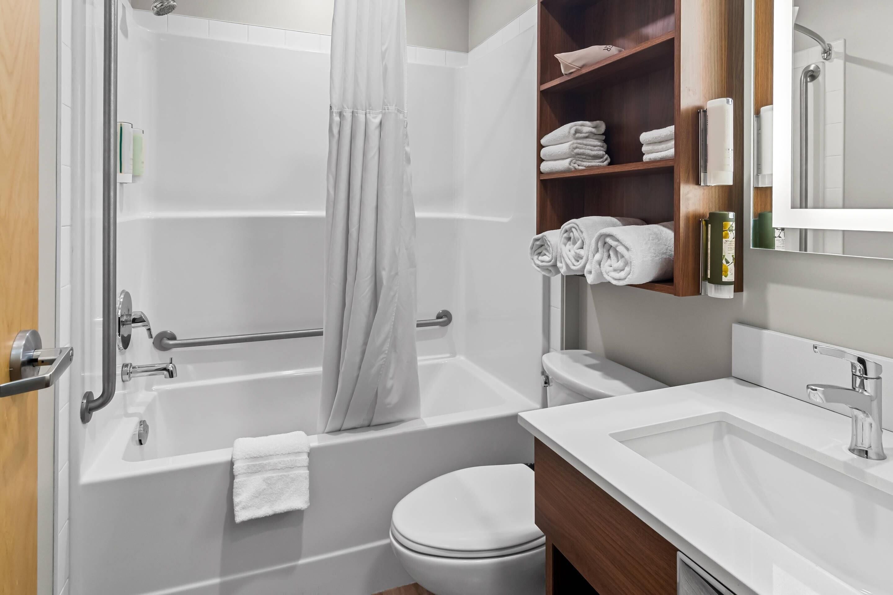 Un baño moderno con bañera, inodoro y lavabo. Hay toallas y artículos de aseo en estantes.