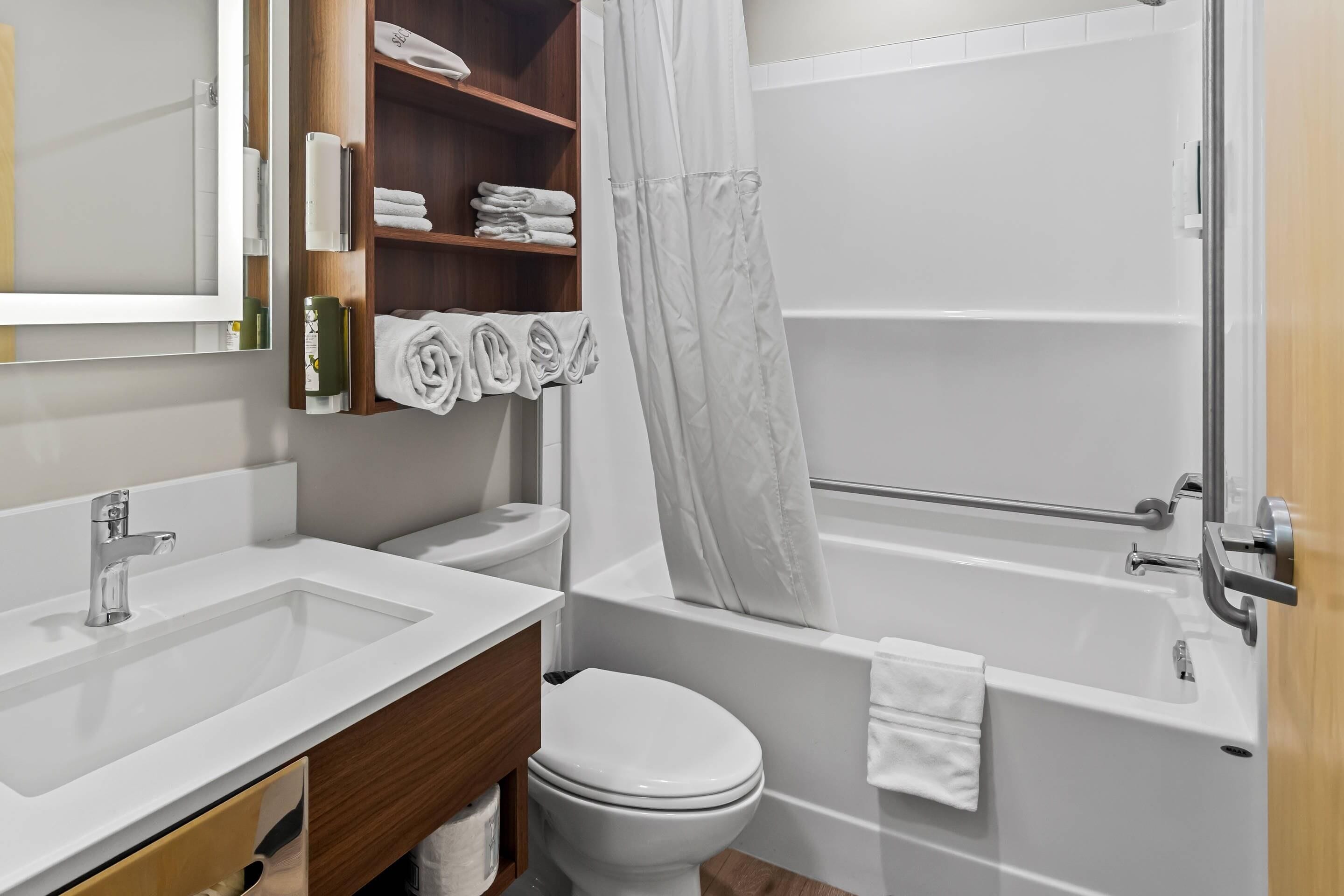 A modern bathroom with a white sink, toilet, and bathtub. There is a wooden shelf with towels and toiletries.
