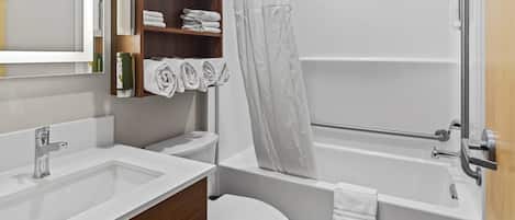 Un baño moderno con lavamanos, inodoro y bañera blancos. Hay un estante de madera con toallas y artículos de aseo.