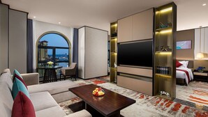 Habitación de hotel moderna con sofá, mesa de comedor con fruta, televisor y vistas a la ciudad.