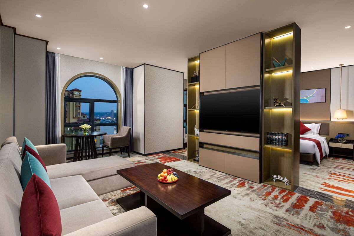 Une chambre d’hôtel moderne avec un canapé, une table à manger avec des fruits, une télévision et une vue sur la ville.
