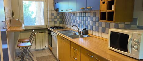 Una cocina compacta con un lavamanos, estufa y microondas. Hay una ventana con persianas, un radiador y un juego de armarios de madera.