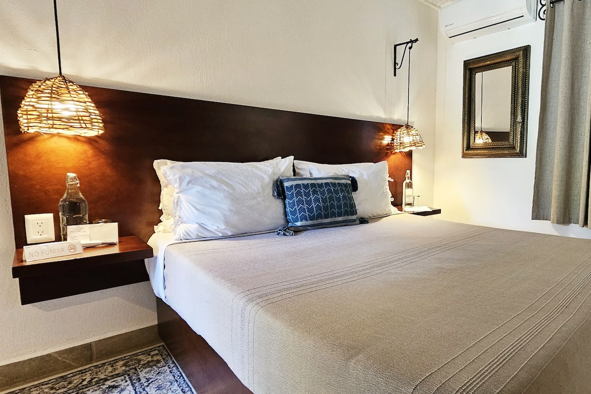 A neatly made bed with white and patterned pillows, a bedside table with a bottle and a glass, and a wall-mounted mirror.