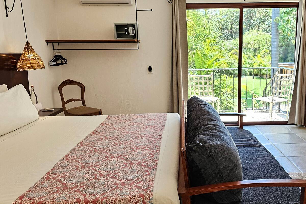 A bedroom with a bed, a patterned bedspread, a wooden chair, and a balcony with a view of greenery.