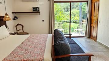 Quarto com cama, colcha estampada, cadeira de madeira e varanda com vista para vegetação.