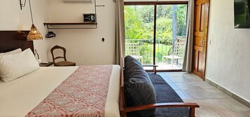 Quarto com cama, colcha estampada, cadeira de madeira e varanda com vista para vegetação.
