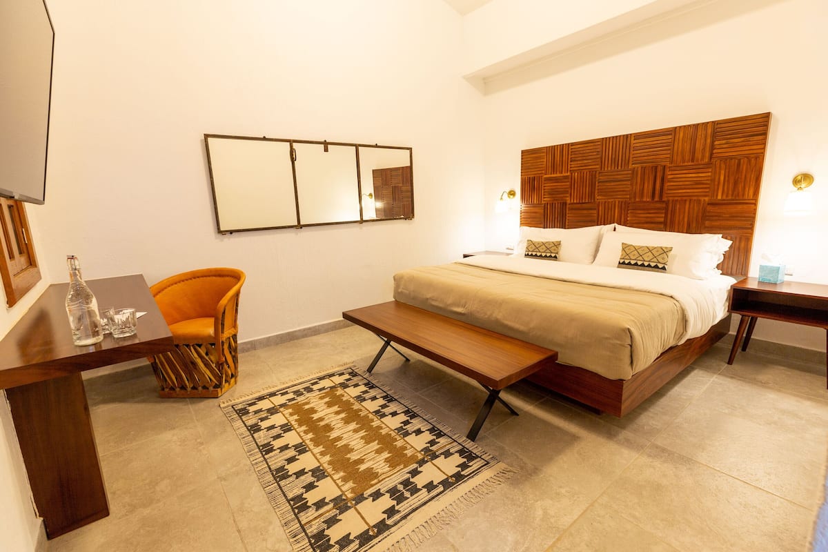 A bedroom with a bed, a wooden headboard, a bench, a chair, a bedside table, a mirror, and a rug.