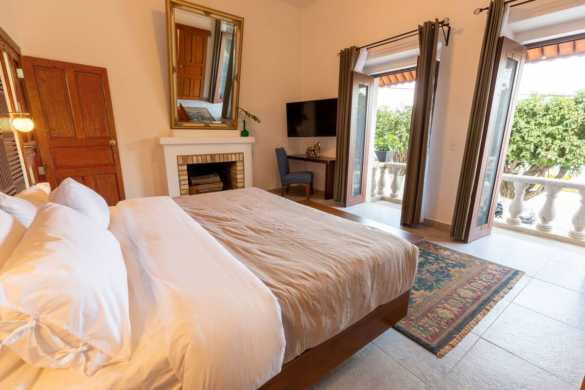 A bedroom with a large bed, a fireplace, a television, and a balcony with curtains.