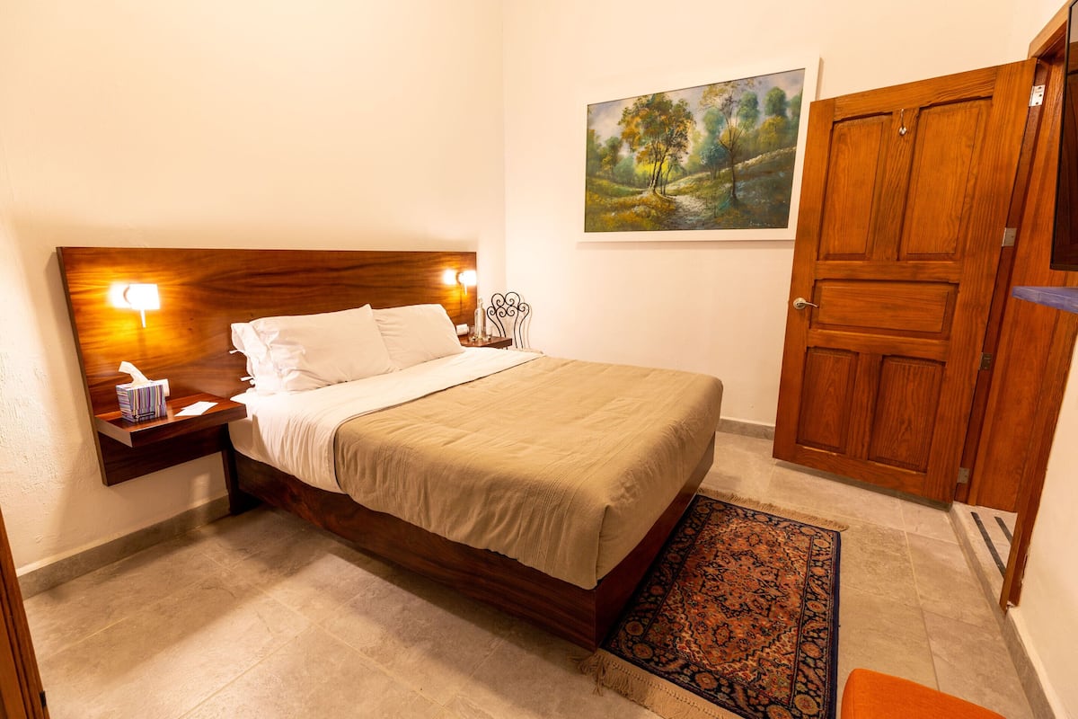 A neatly arranged bedroom with a wooden bed, a nightstand with a lamp, a painting on the wall, and a wooden door.