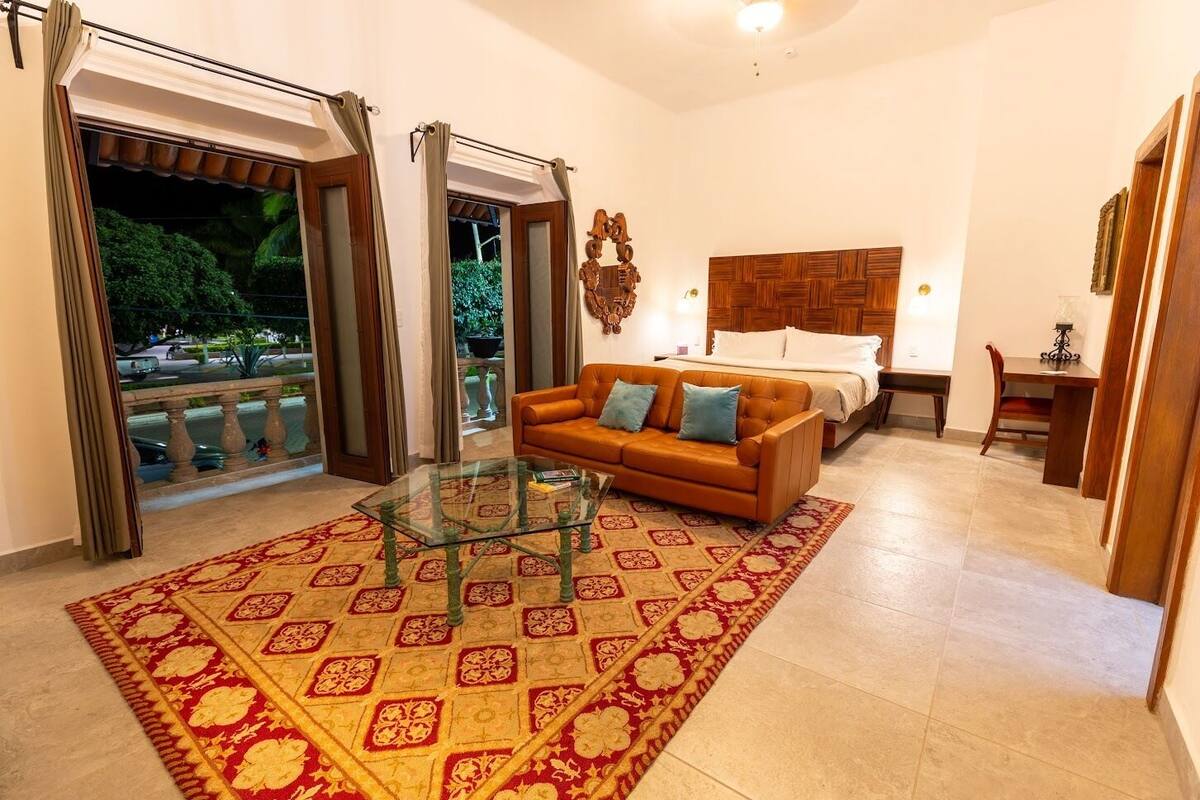 A hotel room with a bed, a sofa, a glass coffee table, and a view of the outdoors.
