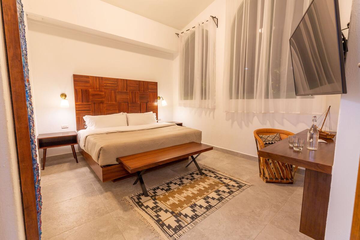 A bedroom with a wooden headboard, a bed with white linens, a bedside table with a lamp, a bench at the foot of the bed, a woven chair, a small table with a bottle and glass, and a TV mounted on the wall.
