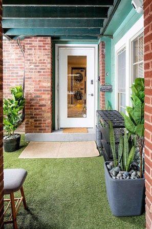Um edifício de tijolos com toldo verde, uma porta com painel de vidro e uma pequena área externa com uma cadeira e uma mesa.