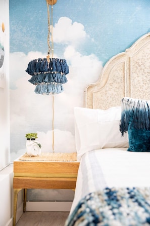 Quarto com cama, mesa de cabeceira, abajur com franjas azuis penduradas e um mural de nuvens na parede.