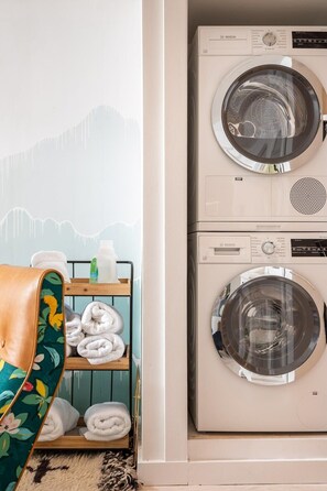 A laundry room with two stacked Bosch washing machines, a wooden shelf with folded towels, and a chair with a floral pattern.