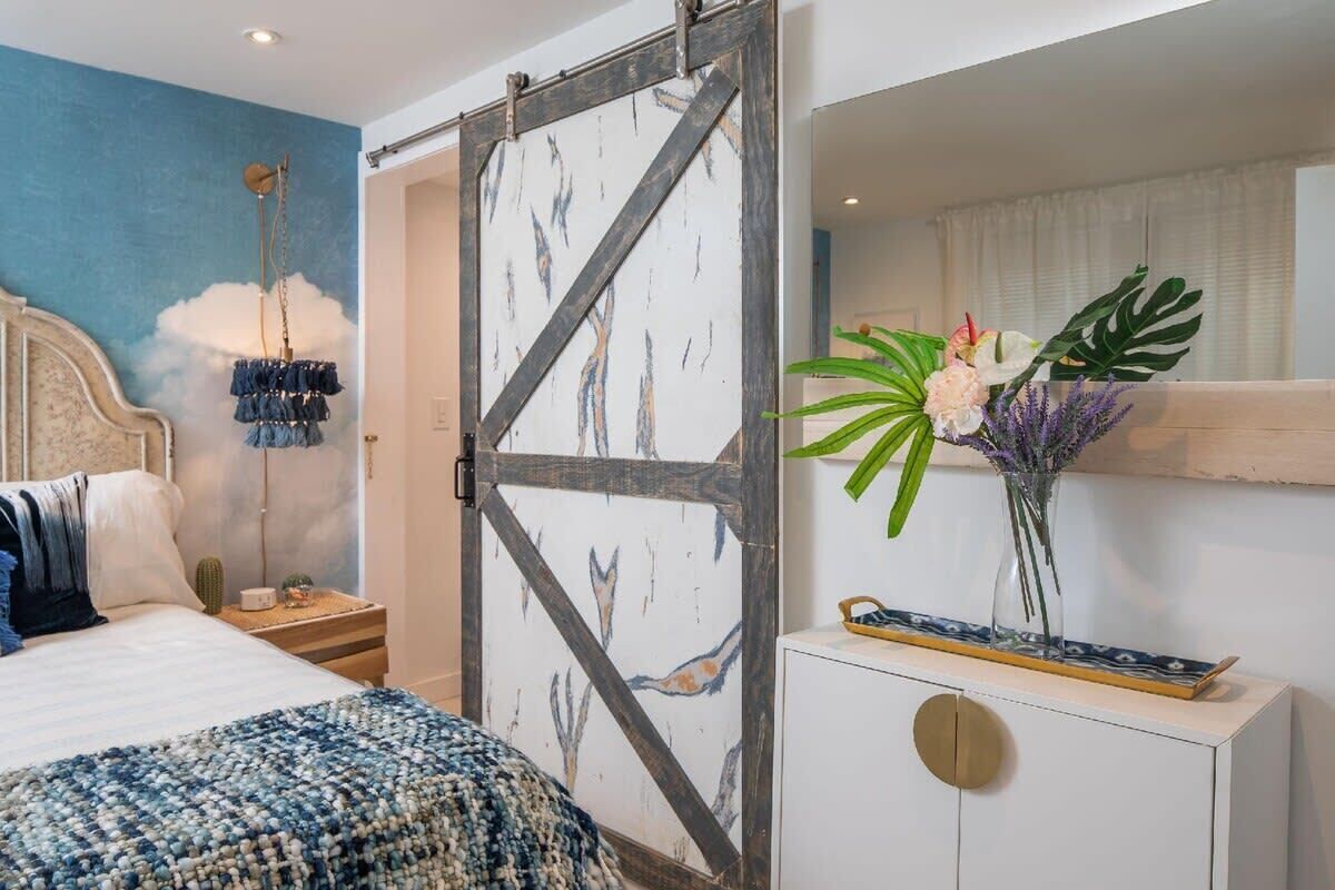 A bedroom with a bed, a nightstand, a dresser, a wall-mounted lamp, and a decorative plant.