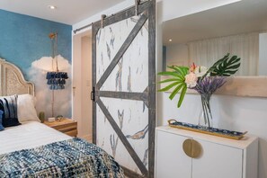 Une chambre à coucher comprenant un lit, une table de chevet, une commode, une lampe fixée au mur et une plante décorative.