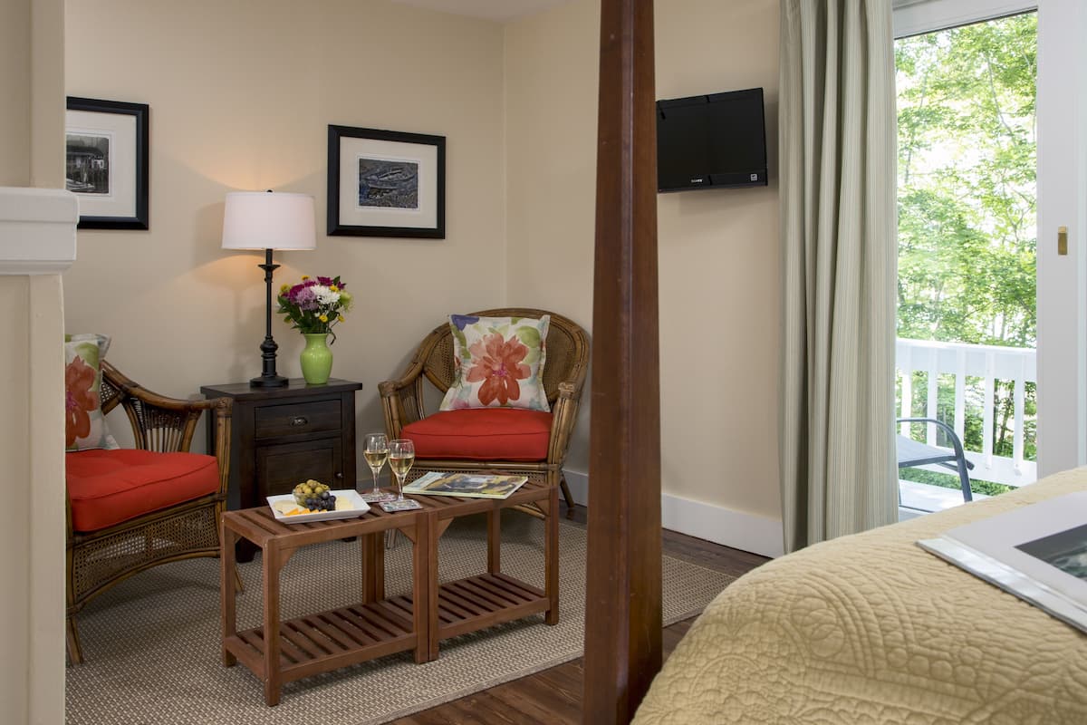 A hotel room with a bed, two wicker chairs with red cushions, a small wooden table with a magazine and glasses, a vase with flowers, and a TV on the wall.