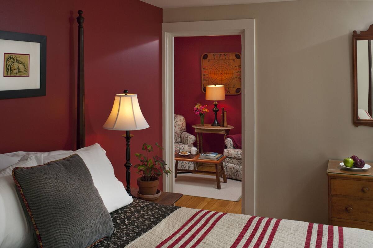 A bedroom with a bed, a nightstand, a lamp, a chair, a small table with a vase of flowers, and a wall clock.