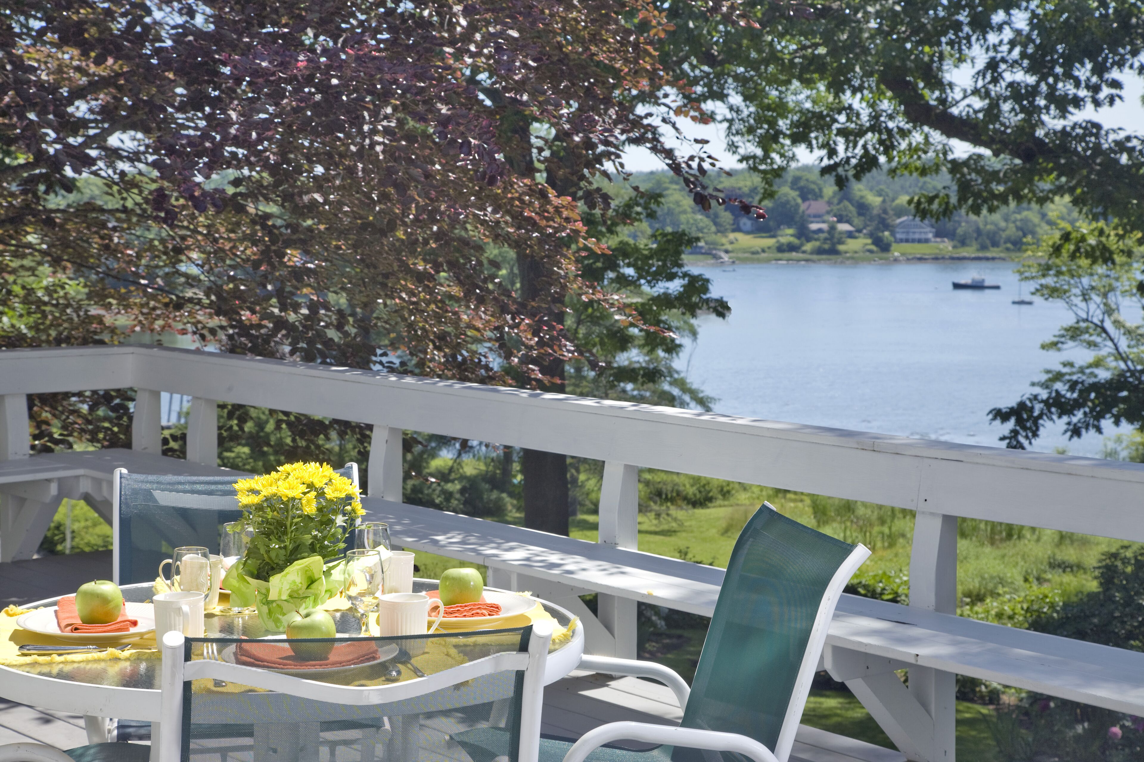 A glass table with a white chair, a glass vase with yellow flowers, and a plate with an apple and a cup on a deck overlooking a lake with boats.