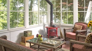 A cozy room with wicker furniture, a glass table, a red stove, and large windows with a view of trees. - Newcastle Inn (Newcastle)