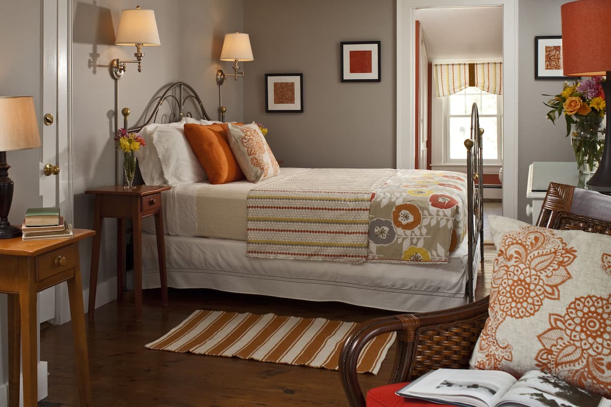A bedroom with a bed, bedside tables, a nightstand, a chair, a lamp, a window with curtains, and a wall with framed pictures.
