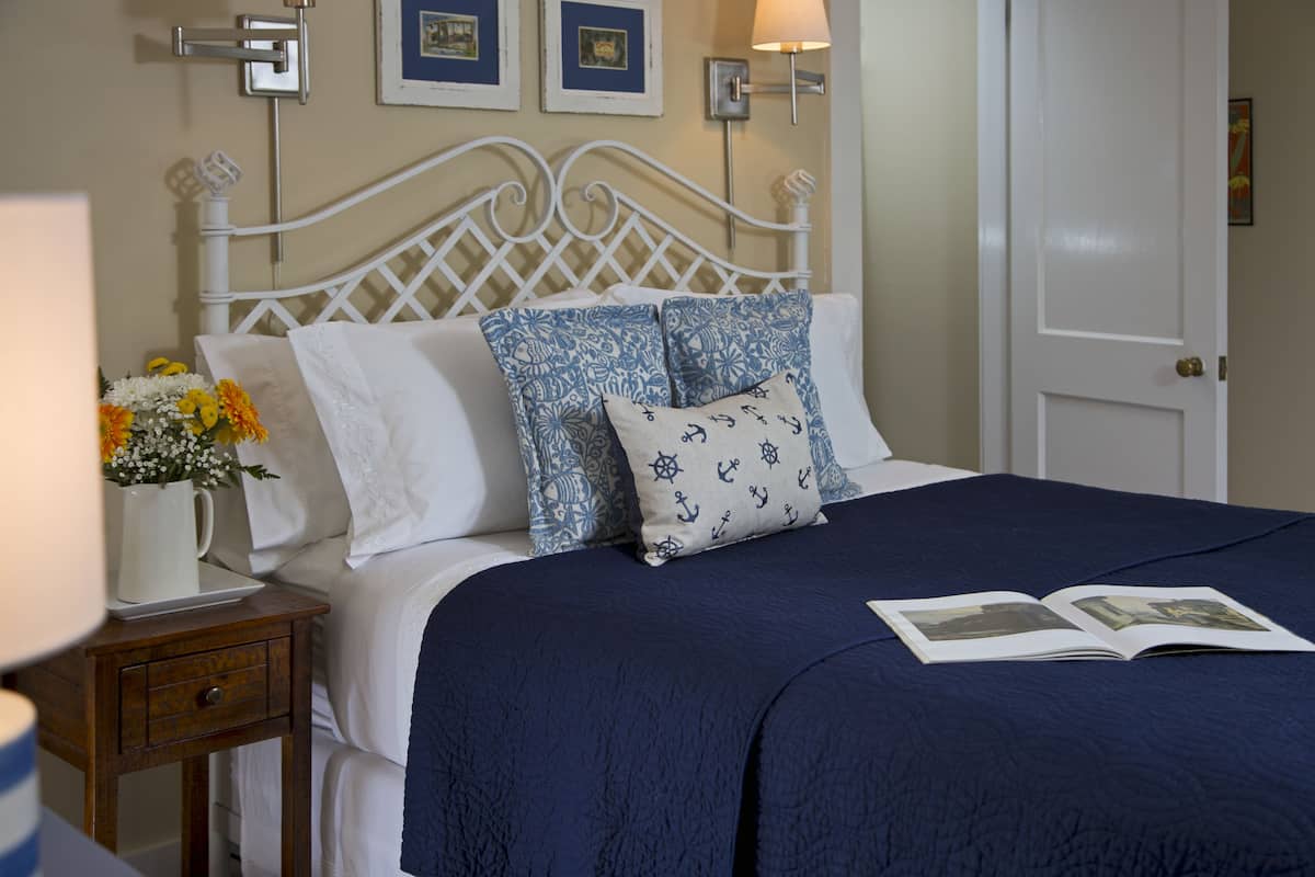 A neatly made bed with a blue bedspread and decorative pillows, a bedside table with a vase of flowers, and framed pictures on the wall.