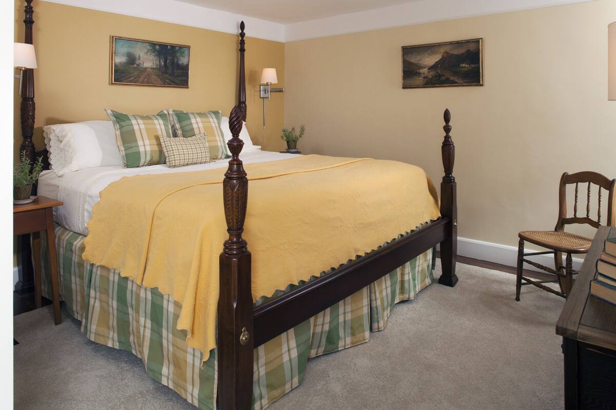 A neatly arranged bedroom with a four-poster bed, a bedside table, a chair, and two paintings on the walls.