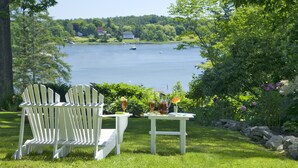 A lakeside view with two Adirondack chairs and a table with drinks, surrounded by greenery and a house in the background. - Newcastle Inn (Newcastle)