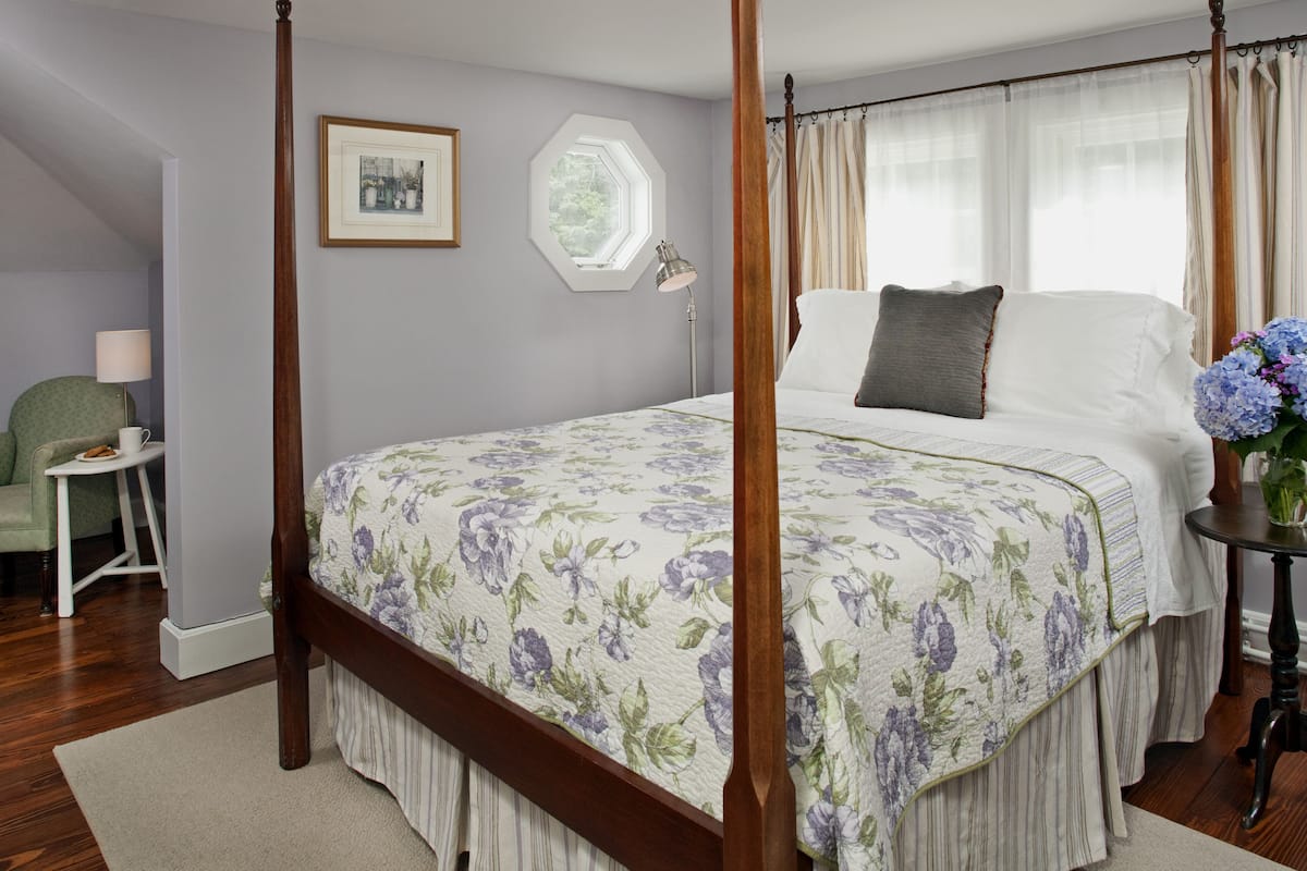 A four-poster bed with a floral bedspread, a bedside table with a lamp, and a chair in a bedroom with a view of the outdoors.