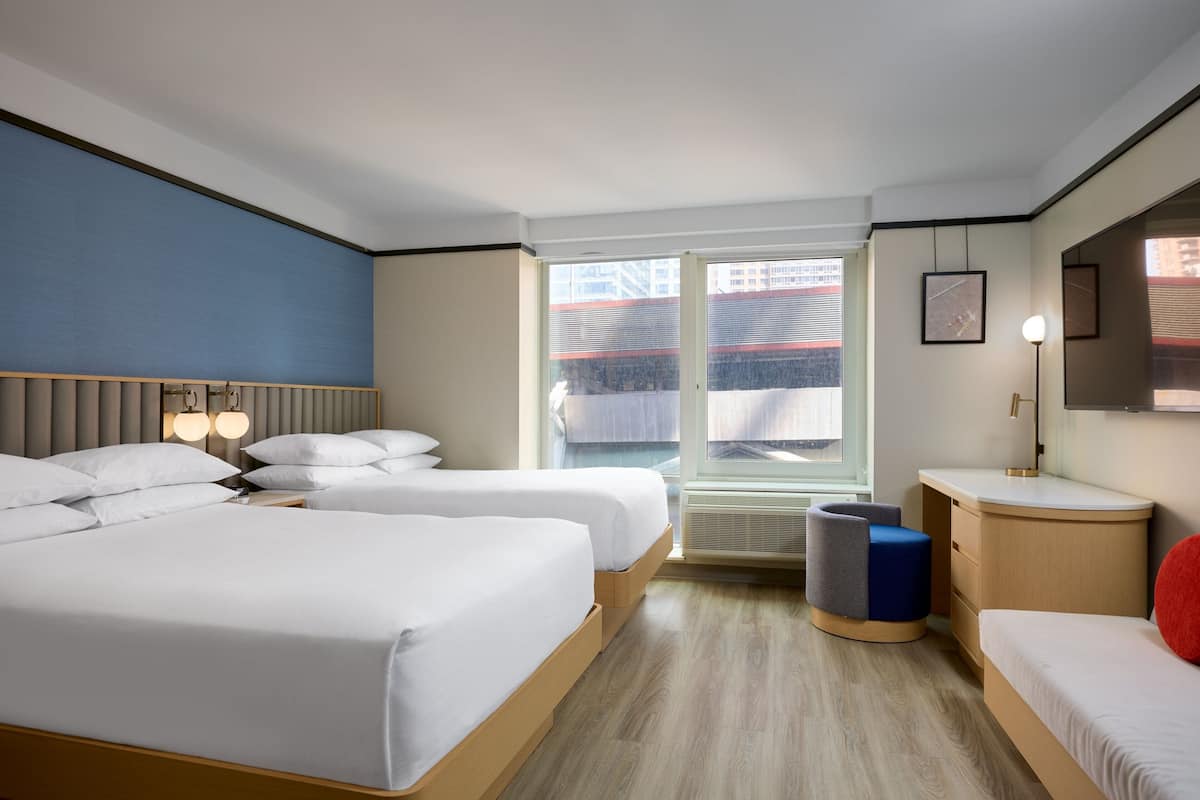 A hotel room with two beds, a small desk, a chair, a window with a view of buildings, and a wall-mounted lamp.