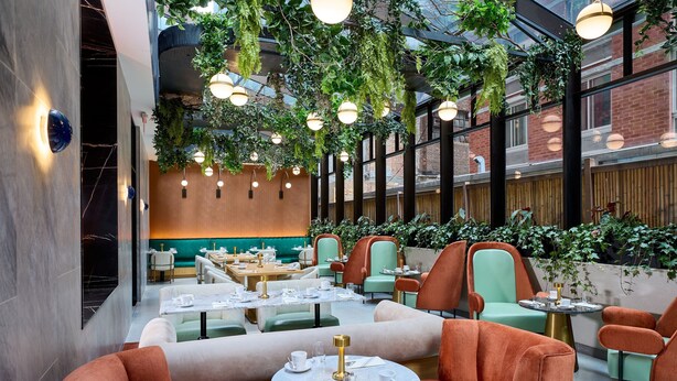Un restaurant avec des tables rondes et des sièges rembourrés, des plantes d’intérieur et des luminaires suspendus.