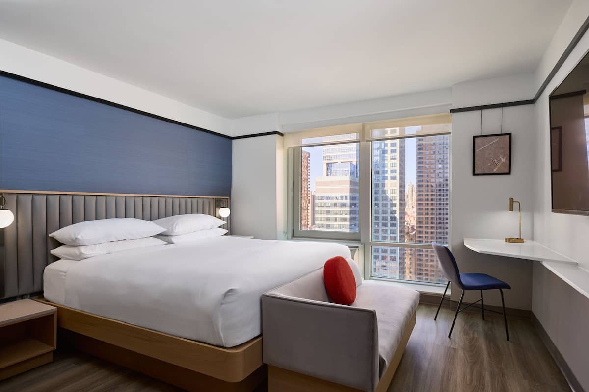 A modern hotel room with a large bed, a desk, and a chair. There is a window with a city view.