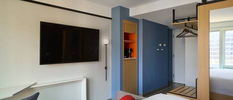 Een moderne hotelkamer met een bed, een bureau, een stoel, een kledingkast en een aan de muur bevestigde televisie.