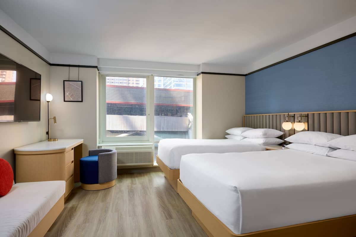 A hotel room with two beds, a desk, a chair, a lamp, and a window with a view of buildings.