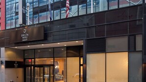 Une entrée d’hôtel moderne avec une façade en verre, une enseigne bien visible indiquant « Delta Hotels », et des drapeaux à l’extérieur.
