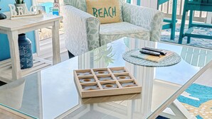 Une table basse avec un plateau en verre et un plateau en bois, un canapé agrémenté d’un coussin orné du mot « PLAGE », et un tapis bleu à motifs.