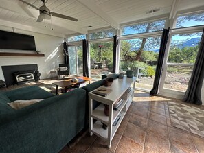 A modern living room with a sectional sofa, a fireplace, a ceiling fan, and large windows offering a view of the outdoors. - Chic 2B2b Bungalow w/ studio 3 mi to Sequoia NP - Dog Friendly! (Three Rivers)