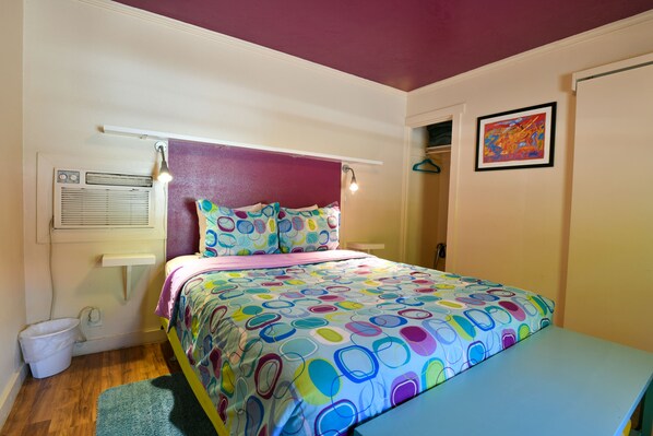 Un dormitorio con una colcha colorida, una mesita de noche, un calentador de pared y un pequeño cubo de basura.