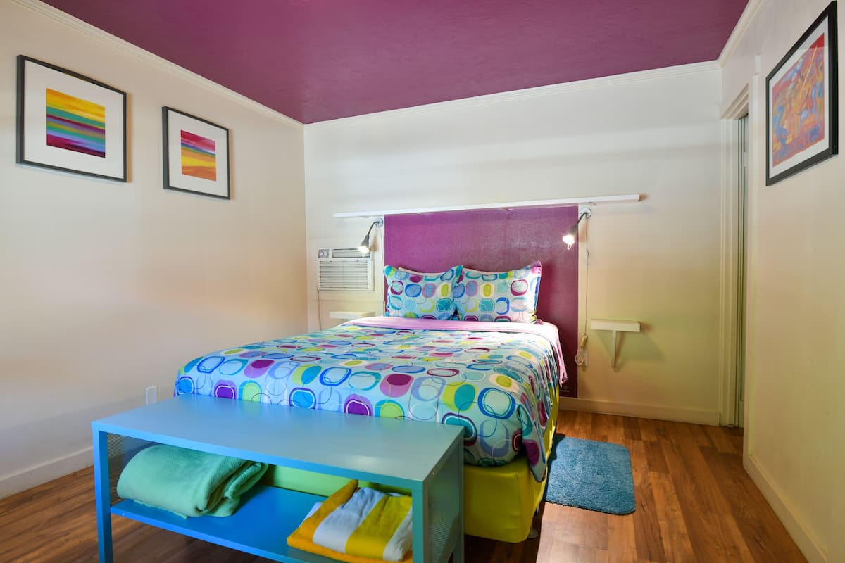 A bedroom with a bed, a small table with towels, a window, and a door.