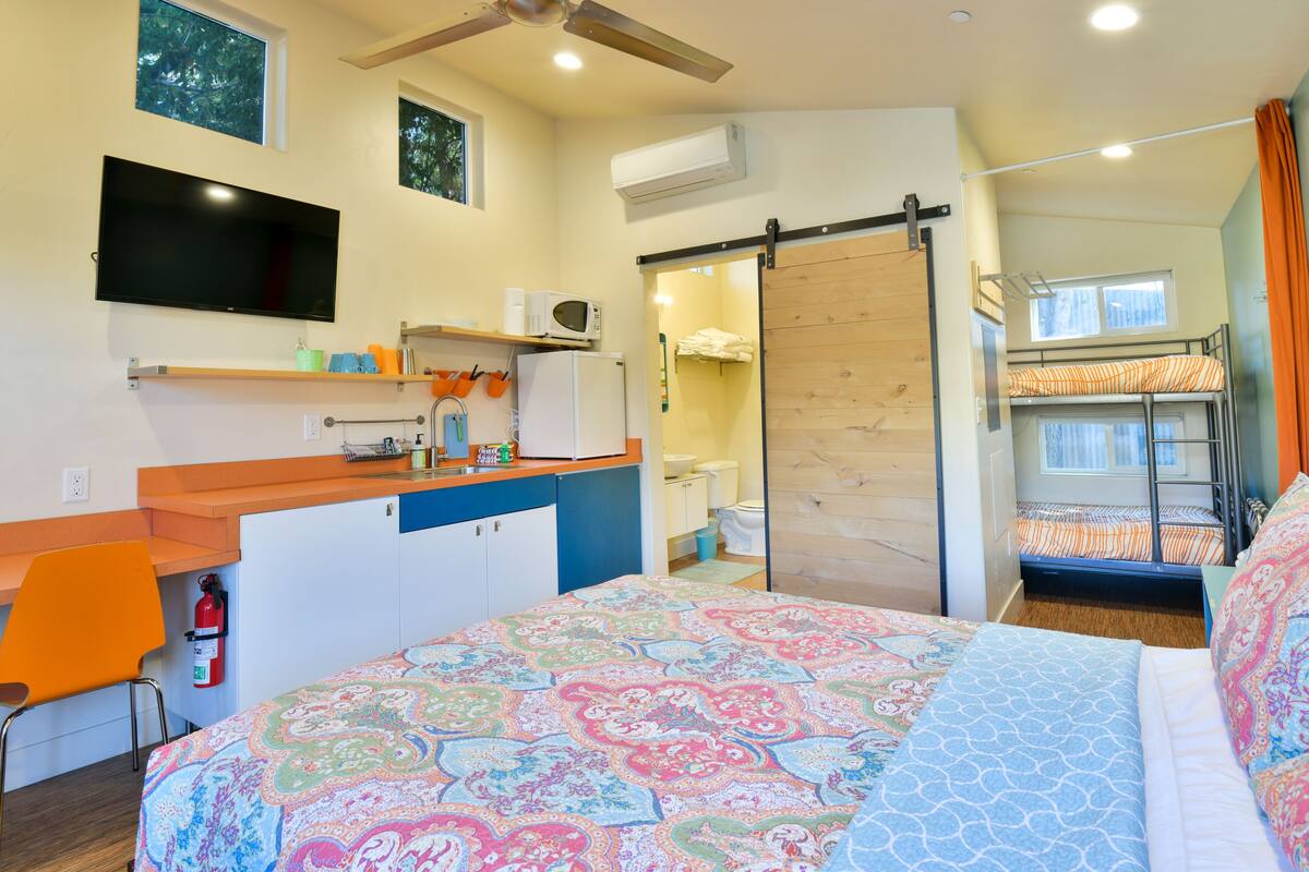Quarto de hotel pequeno com cama, escrivaninha, cadeira, televisão, micro-ondas e armário com porta deslizante.